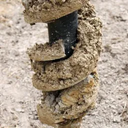 Wiertło świdrowe do gruntu, pokryte wilgotną ziemią, w trakcie wiercenia otworu w ziemi.