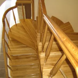 Spiralne schody drewniane z balustradą, widok z góry, jasne drewno, polakierowane, widoczne detale stopni i poręczy.