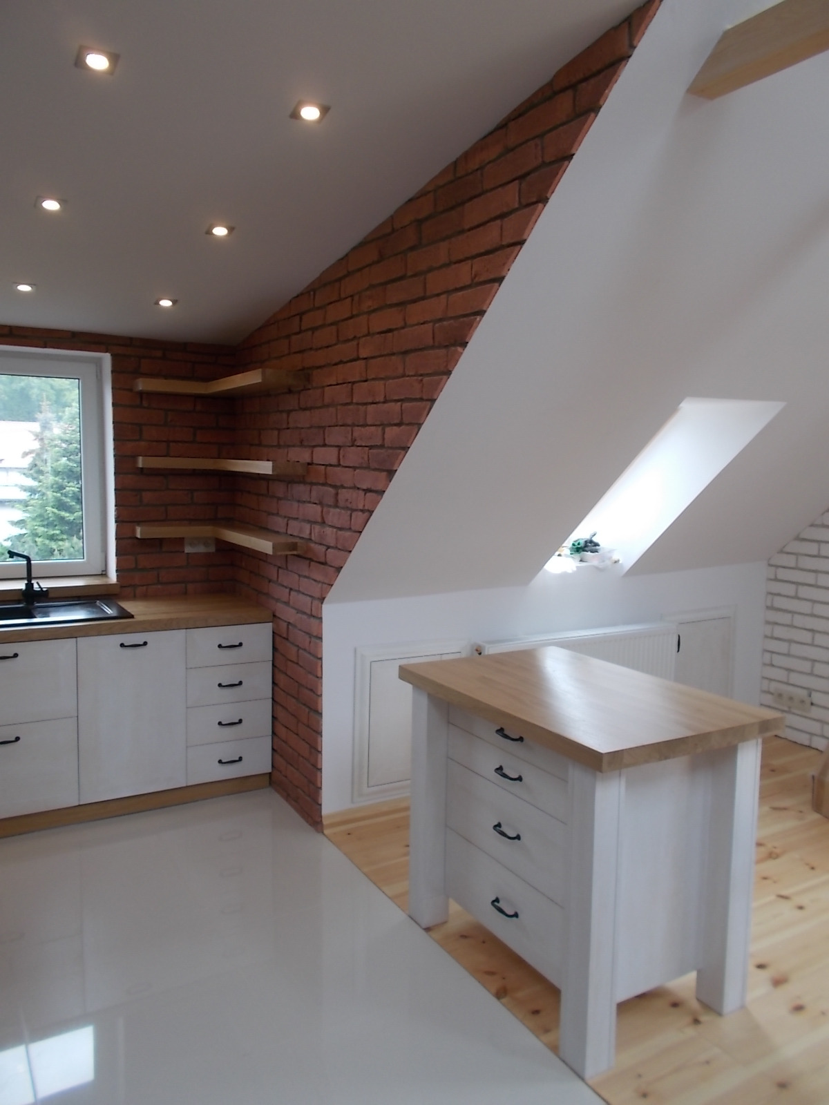 Jasna kuchnia na poddaszu z ceglaną ścianą, oświetleniem punktowym i drewnianymi blatami, widoczna wyspa kuchenna z szufladami i szafki pod oknem.
