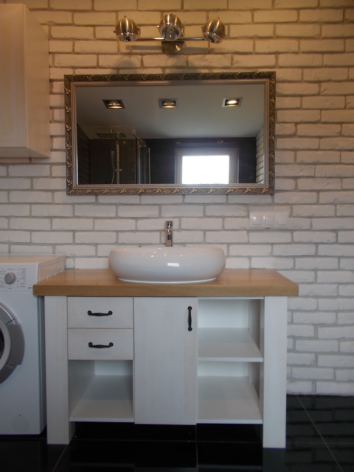 Biała umywalka nablatowa w owalnym kształcie, osadzona na drewnianym blacie szafki łazienkowej z białymi frontami i czarnymi uchwytami, na tle ściany z białej cegły, z lustrem w ozdobnej ramie...
