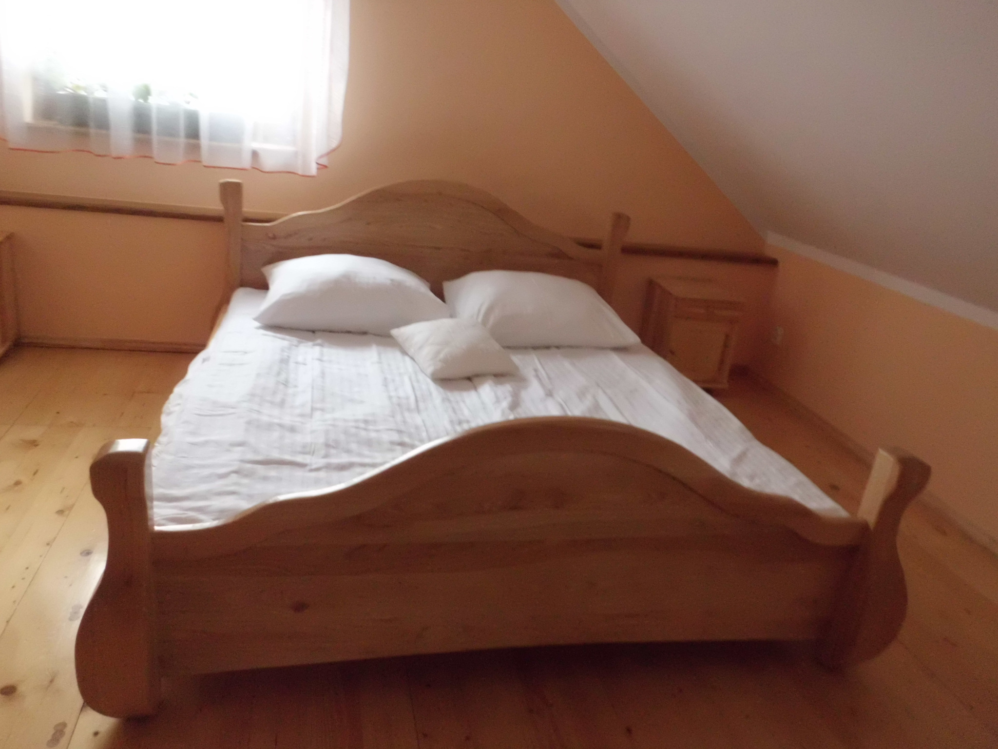 Drewniane łóżko z białymi poduszkami w pokoju na poddaszu z oknem i małą szafką nocną.