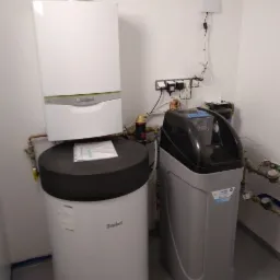 Kompaktowa kotłownia w pomieszczeniu technicznym: wiszący kocioł gazowy Vaillant ecoCompact nad zasobnikiem wody 150l, obok stacja uzdatniania wody z szarą obudową, instalacja rurowa na ścianie.