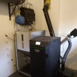 Zainstalowana kotłownia pelletowa z czarnym kotłem Kostrzewy EEI PELLETS 10kW z zielonymi akcentami, widoczny zasobnik peletu i elementy instalacji grzewczej w pomieszczeniu kotłowni.