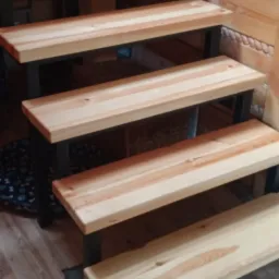 Schody drewniane z metalowymi wspornikami w drewnianym domu, widoczne słoje drewna i fragment podłogi.