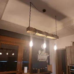 Drewniana lampa wisząca z trzema żarówkami Edison na łańcuchach, oświetlająca blat kuchenny z dekoracjami w stylu rustykalnym.