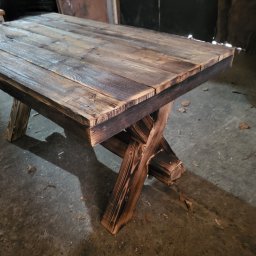 Sewostyle Meble na wymiar altanki spawanie - Stolik kawowy z blatem z desek o wyraźnej strukturze drewna, wsparty na nogach w kształcie litery X, wykonanych z opalanych belek. Surowy, rustykalny styl.