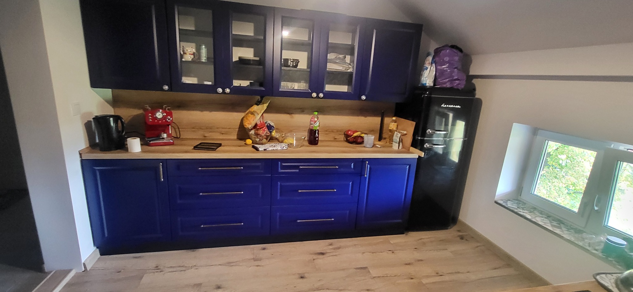 Niebieska kuchnia z drewnianym blatem pod skosem dachu, wyposażona w czarny retro lodówkę, ekspres do kawy i szklane szafki wiszące.