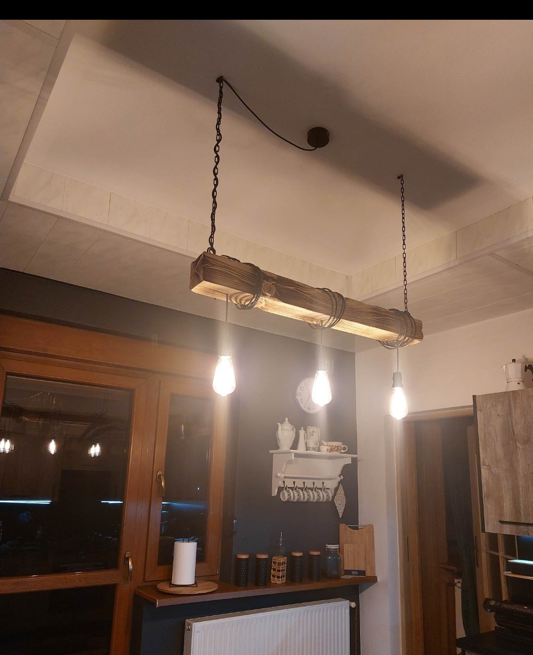 Drewniana lampa wisząca z trzema żarówkami Edison na łańcuchach, oświetlająca blat kuchenny z dekoracjami w stylu rustykalnym.