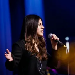 Kobieta z mikrofonem śpiewa na scenie w blasku niebieskich świateł, widoczny profil twarzy i rozpuszczone brązowe włosy.