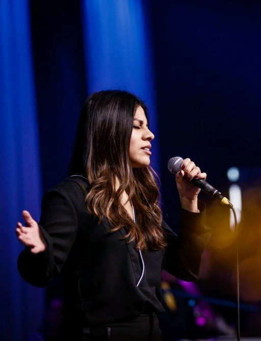 Kobieta z mikrofonem śpiewa na scenie w blasku niebieskich świateł, widoczny profil twarzy i rozpuszczone brązowe włosy.