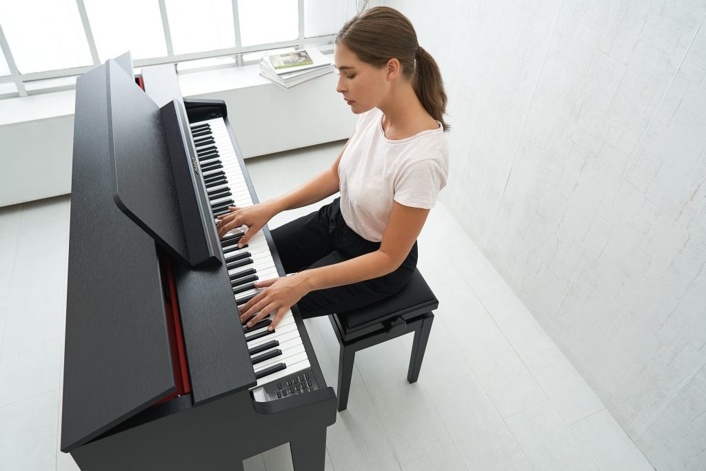 Młoda kobieta gra na czarnym pianinie cyfrowym w jasnym, minimalistycznym wnętrzu z oknem i stertą książek w tle.