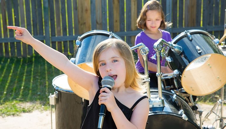 Dziewczynka śpiewa do mikrofonu, wskazując palcem, a w tle druga gra na perkusji. Nauka gry na instrumentach w plenerze, na tle drewnianego płotu.