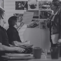 Trzech osób analizujących czarno-białe zdjęcia dowodowe przypięte do ściany w biurze, jeden z mężczyzn trzyma w ręku dokument, a kobieta długopis.