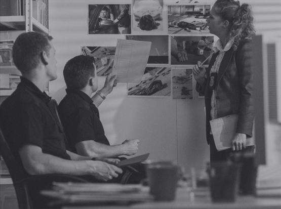 Trzech osób analizujących czarno-białe zdjęcia dowodowe przypięte do ściany w biurze, jeden z mężczyzn trzyma w ręku dokument, a kobieta długopis.