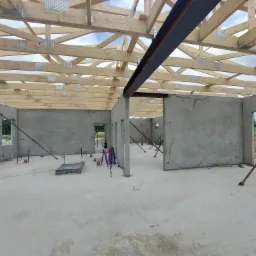 Wnętrze domu murowanego w trakcie budowy, widoczne ściany z betonu komórkowego, drewniana więźba dachowa z metalowymi łącznikami oraz stalowa belka stropowa, podpory stropowe i instalacje w podłodze.