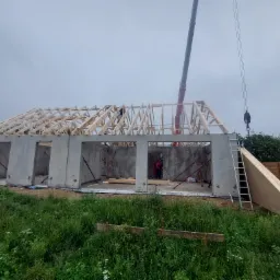 Budowa parterowego domu z prefabrykatów betonowych, widoczna drewniana więźba dachowa montowana przy użyciu dźwigu, krajobraz polny.