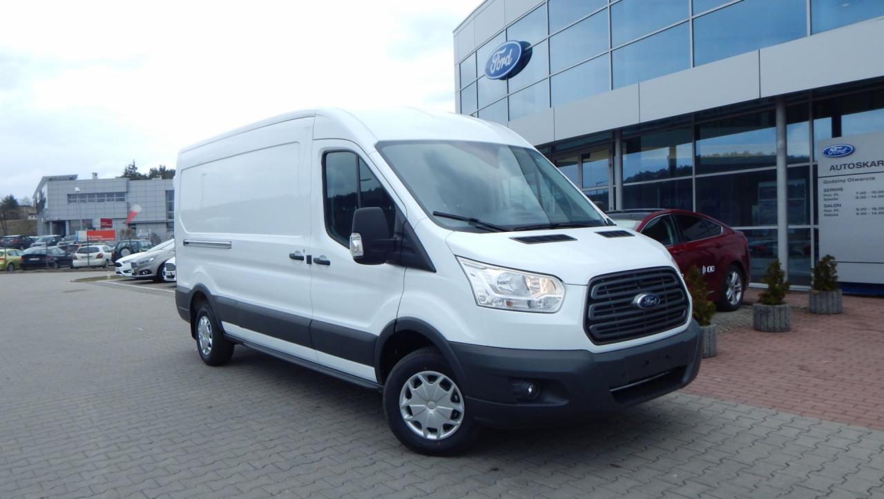 Biały, fabrycznie nowy Ford Transit na placu dealera w Ostrowie Wielkopolskim, widok z przodu, na tle przeszklonego budynku salonu samochodowego.