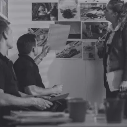 Czarno-białe zdjęcie przedstawia trzech mężczyzn i kobietę analizujących zdjęcia z miejsc zbrodni przypięte do ściany w biurze.