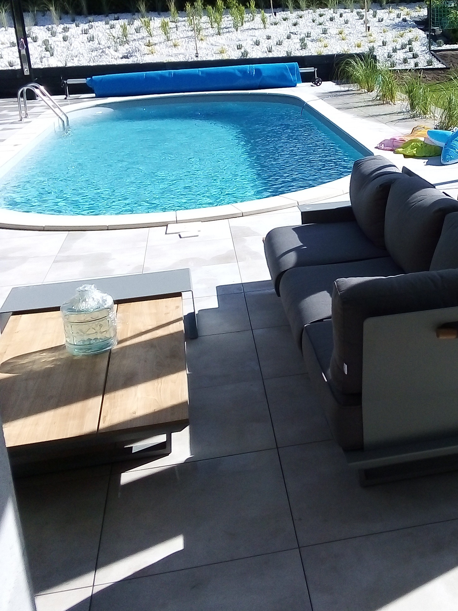 Jasnoszara, nowoczesna sofa ogrodowa na tarasie z płyt gresowych, obok stolik kawowy z szklanym wazonem na tle basenu z błękitną wodą i białymi kamieniami w ogrodzie.