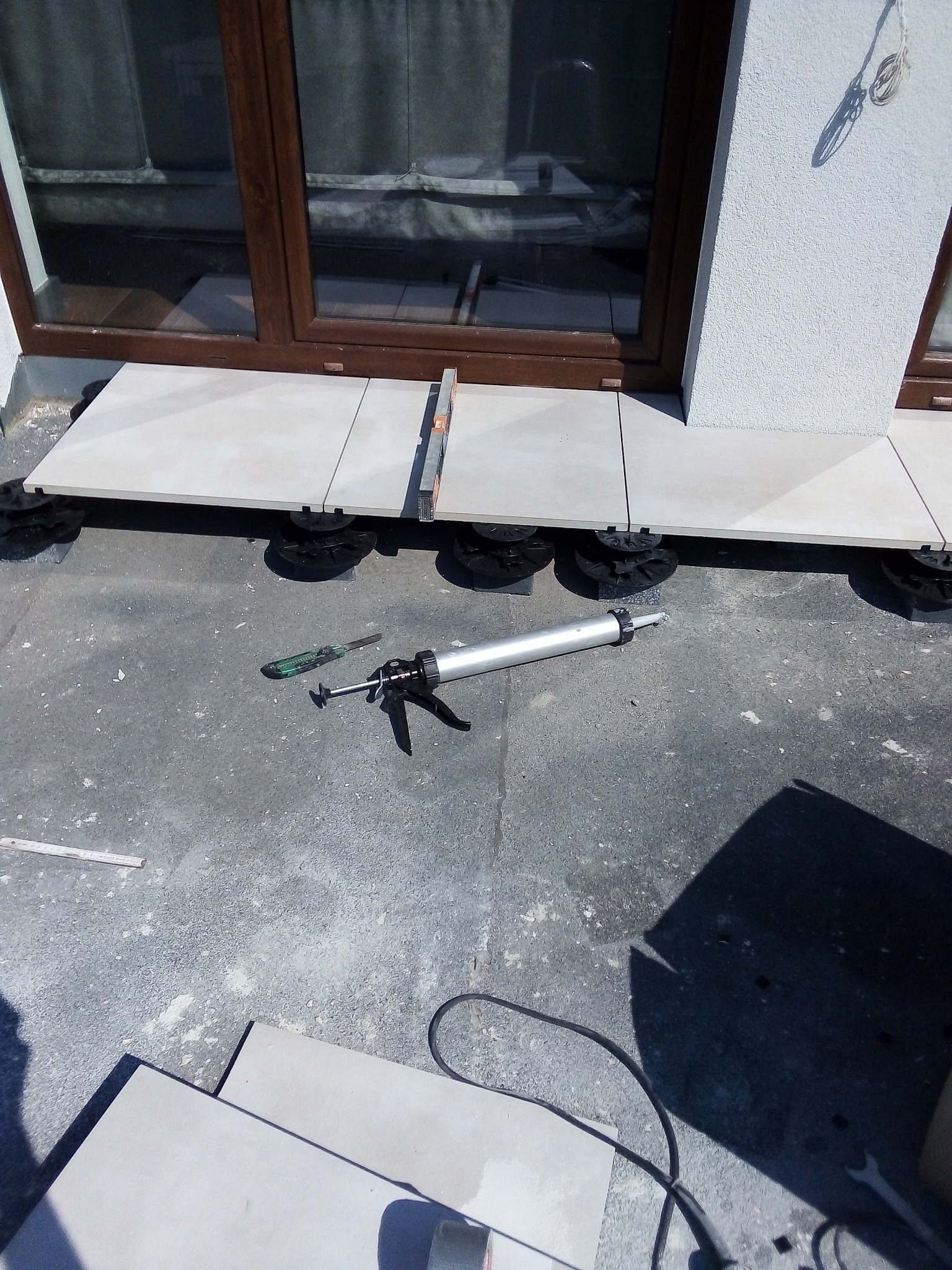 Układanie jasnych płytek na tarasie z użyciem regulowanych wsporników; widoczny pistolet do kleju, nóż i poziomica