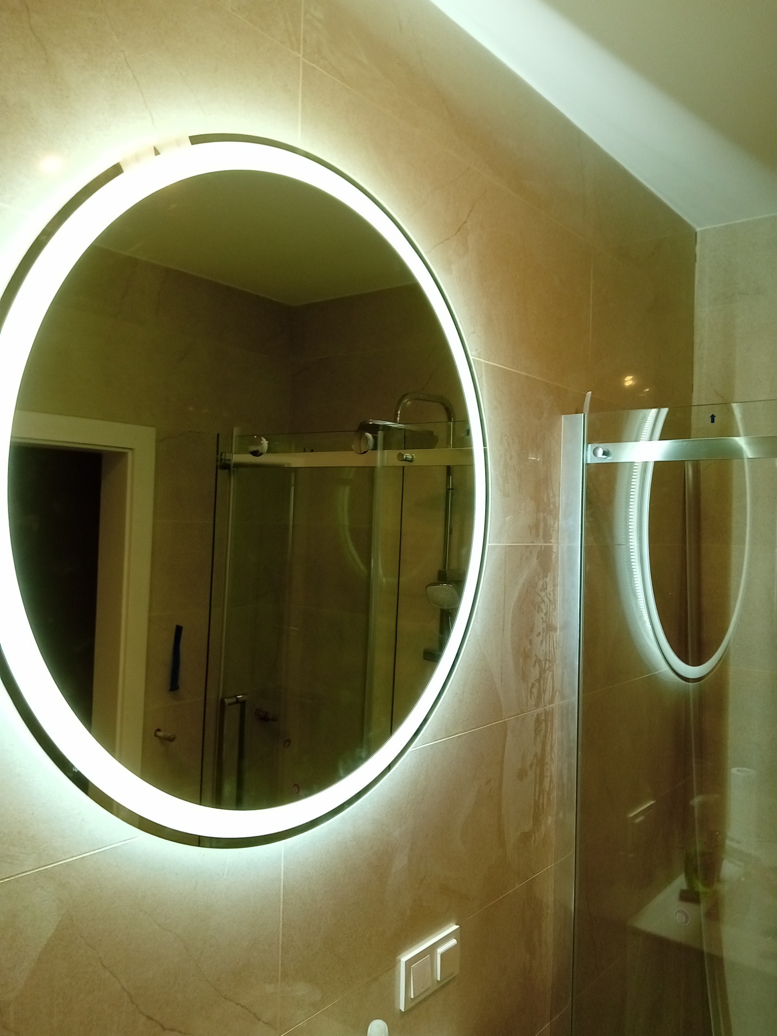 Okrągłe lustro z podświetleniem LED w łazience wyłożonej beżowymi płytkami, odbijające kabinę prysznicową i drugie lustro, widoczne włączniki światła na ścianie.