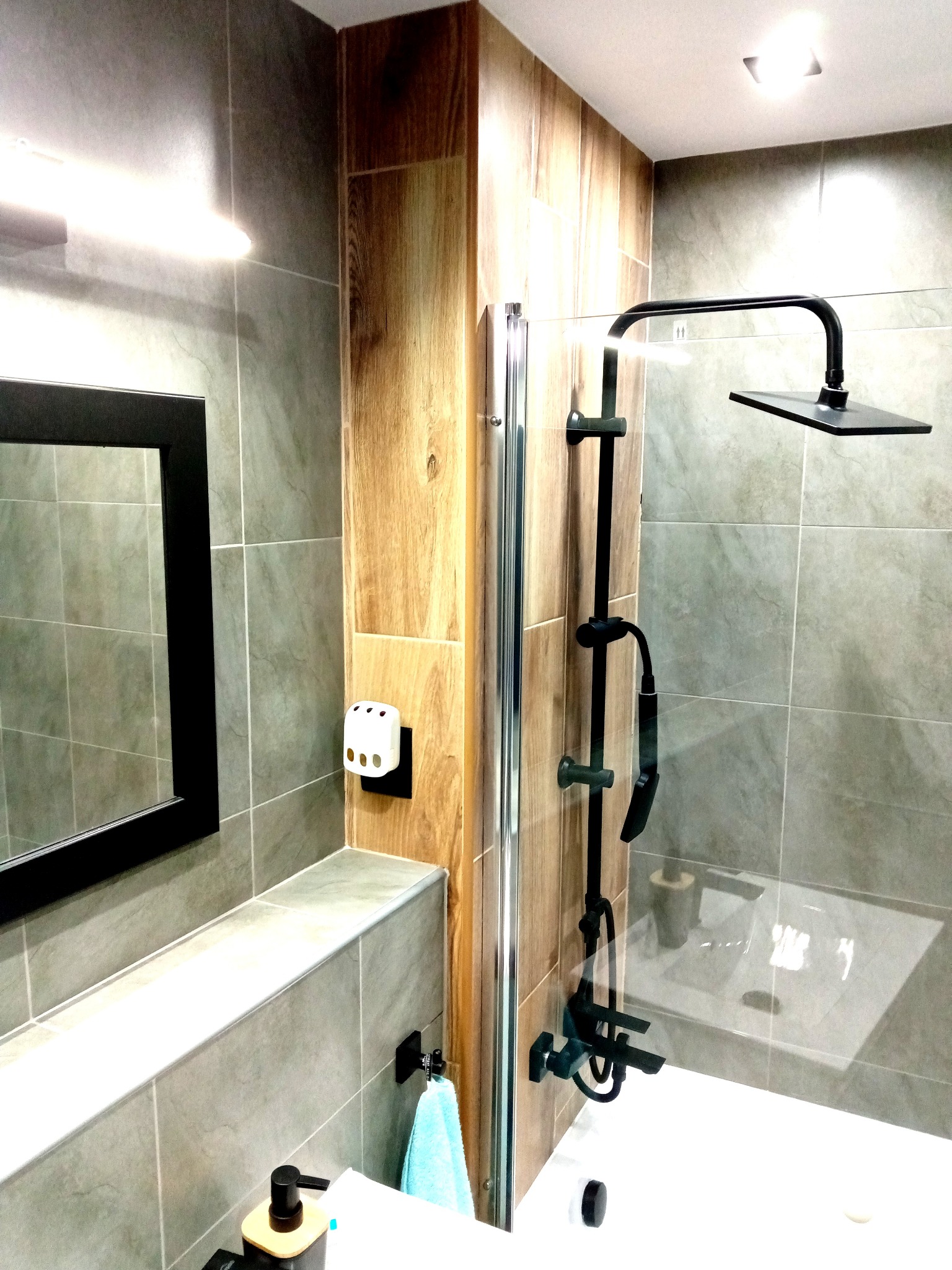 Nowoczesna łazienka z szarymi płytkami, drewnianym akcentem, czarną armaturą i minimalistycznym lustrem w czarnej ramie. Widoczny fragment kabiny prysznicowej z deszczownicą.