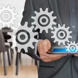 Mężczyzna w szarym garniturze trzyma tablet z interaktywnymi grafikami kół zębatych, symbolizującymi procesy, na tle sali konferencyjnej.