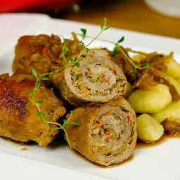 Rolada mięsna przekrojona na pół, faszerowana warzywami, podana z kluskami i sosem cebulowym, udekorowana gałązkami tymianku na białym talerzu.