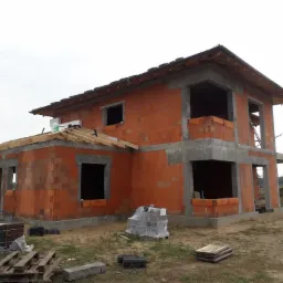 Dwukondygnacyjny dom w trakcie budowy z czerwonej cegły Porotherm, widoczne niezadaszone okna i betonowe elementy konstrukcyjne, na ziemi palety z materiałami budowlanymi.