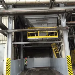 Przemysłowa konstrukcja stalowa z pomalowanymi elementami na szaro i żółto, widoczny pracownik w kasku i kamizelce odblaskowej, stojący obok zabezpieczonej strefy z żółto-czarnymi pasami...