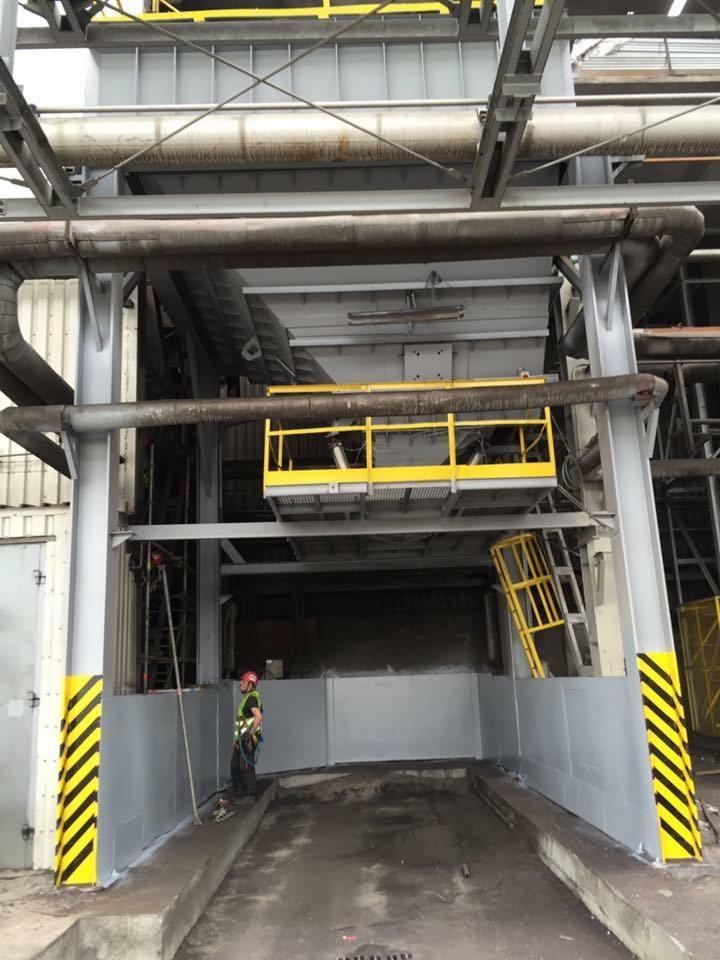 Przemysłowa konstrukcja stalowa z pomalowanymi elementami na szaro i żółto, widoczny pracownik w kasku i kamizelce odblaskowej, stojący obok zabezpieczonej strefy z żółto-czarnymi pasami...