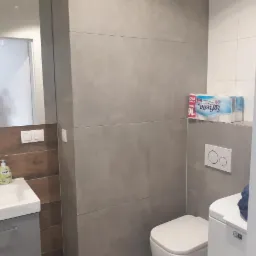 Nowoczesna łazienka z szarymi płytkami wielkoformatowymi, białą armaturą i drewnianym akcentem przy lustrze, uzupełniona o dozownik mydła Isana i zapas papieru toaletowego Softella.