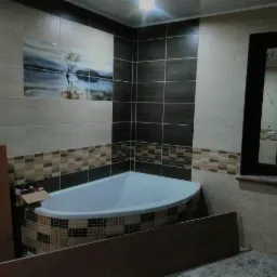 Narożna wanna obudowana mozaiką w łazience z brązowo-beżowymi płytkami i dekoracyjnym panelem z motywem pejzażu.