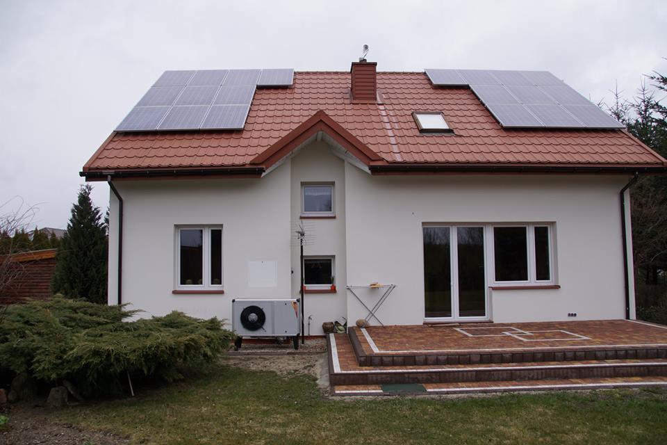 Dom jednorodzinny z zamontowanymi panelami słonecznymi na dachu z czerwonej dachówki, widoczny klimatyzator na zewnątrz budynku, mała antena satelitarna na ścianie.