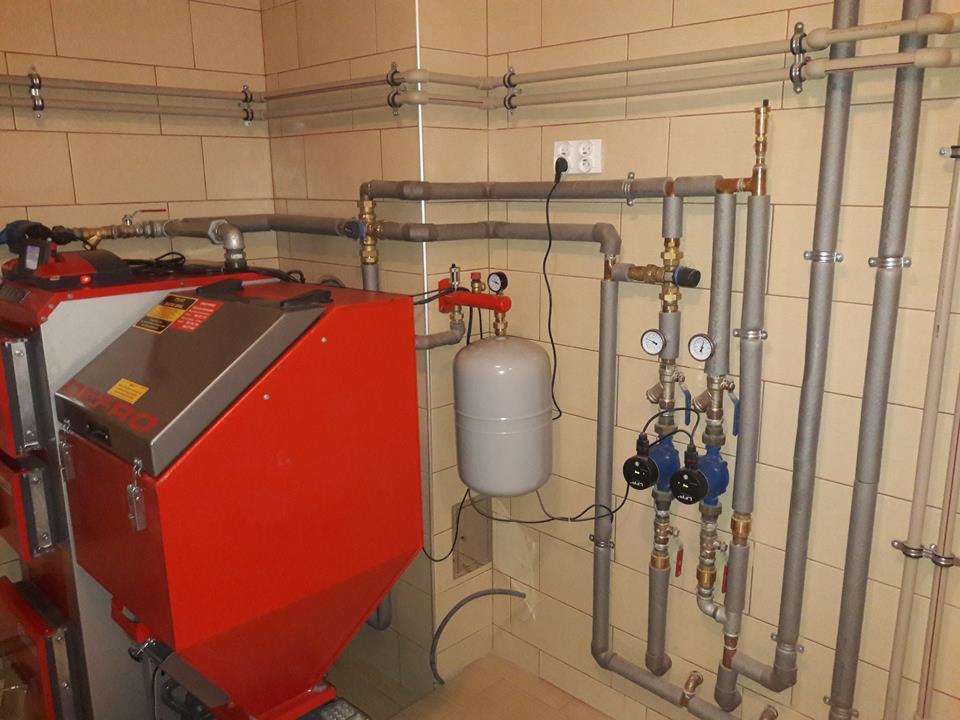 Czerwony piec CO z szarymi rurami instalacji grzewczej, manometry i zbiornik wyrównawczy na tle beżowych płytek.