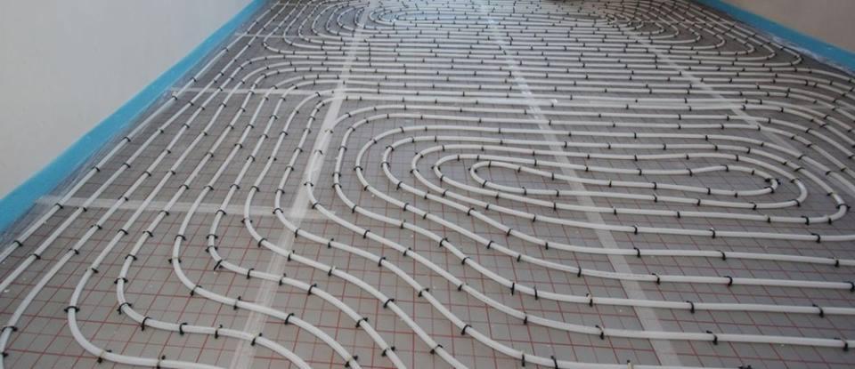 Białe rury systemu ogrzewania podłogowego, mocowane czarnymi klipsami do siatki na szarym podłożu, widok z góry