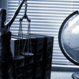 Statua Temidy z wagą sprawiedliwości, obok globus i książki o prawie, na tle żaluzji okiennych w odcieniach niebieskiego.