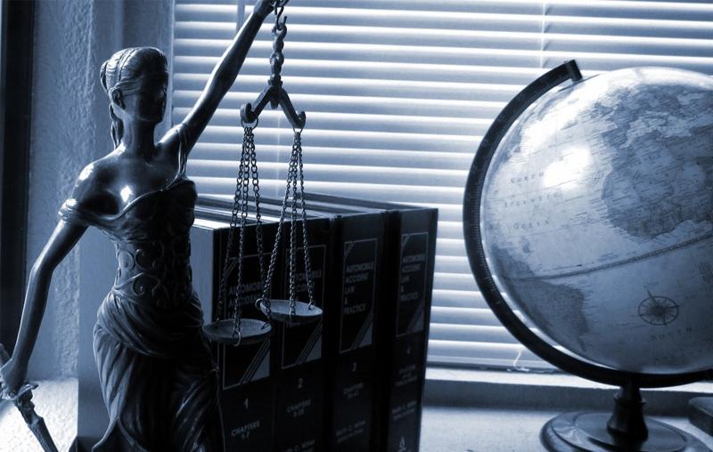 Statua Temidy z wagą sprawiedliwości, obok globus i książki o prawie, na tle żaluzji okiennych w odcieniach niebieskiego.