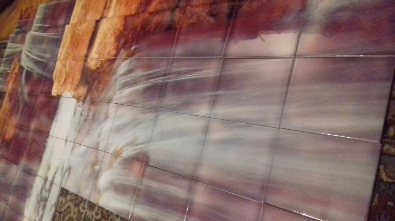 Ściana wyłożona błyszczącymi, kwadratowymi płytkami z motywem wodospadu i skał w odcieniach brązu, bieli i fioletu, widoczna z boku na tle wzorzystego dywanu.