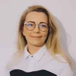 Portret uśmiechniętej blondynki w okularach, ubrana w białą koszulę z czarnym wykończeniem, na jasnym tle.