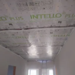 Montaż membrany paroizolacyjnej Intello Plus na poddaszu, widoczne detale łączeń i nadruki na membranie, w tle drabina oparta o ścianę.