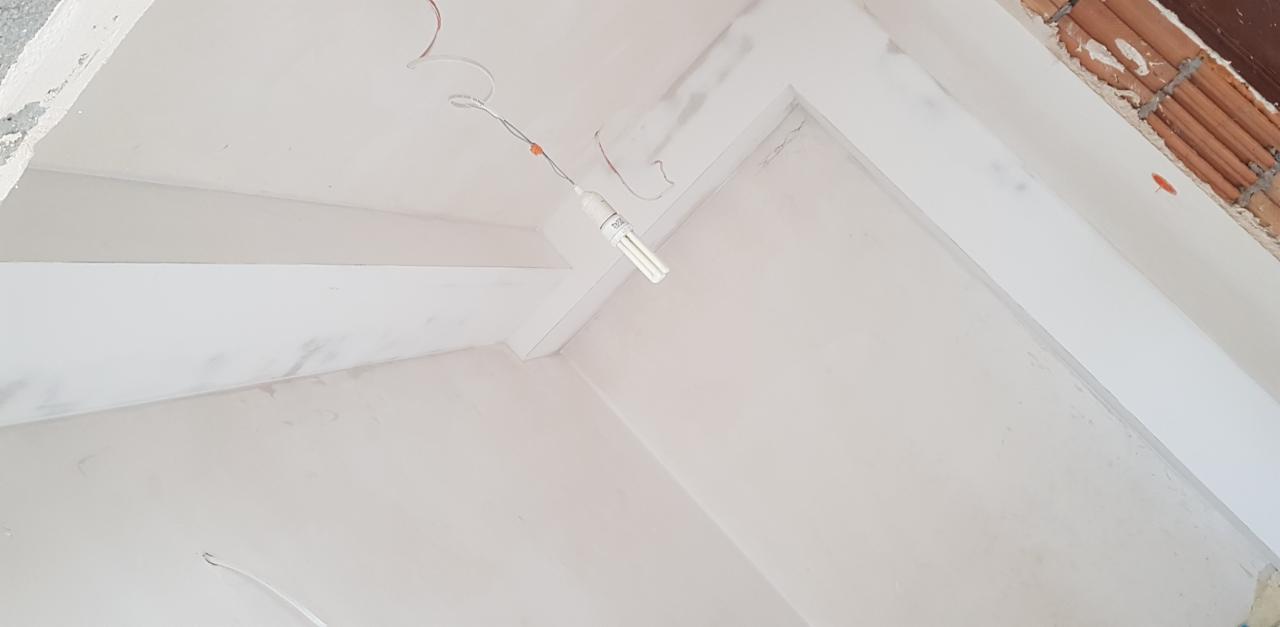 Wnętrze w trakcie prac wykończeniowych, widoczne ściany i sufit po szpachlowaniu, surowy stan z wiszącą żarówką i fragmentem muru z cegieł.