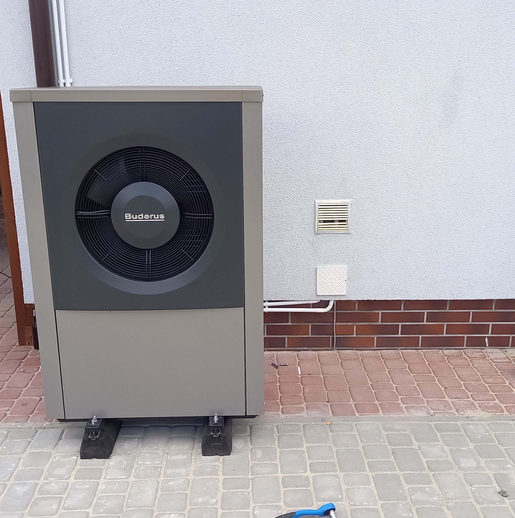 Zewnętrzna jednostka pompy ciepła marki Buderus, zamontowana na czarnych podkładkach, ustawiona przy białej ścianie z ceglaną podmurówką i widoczną kratką wentylacyjną.