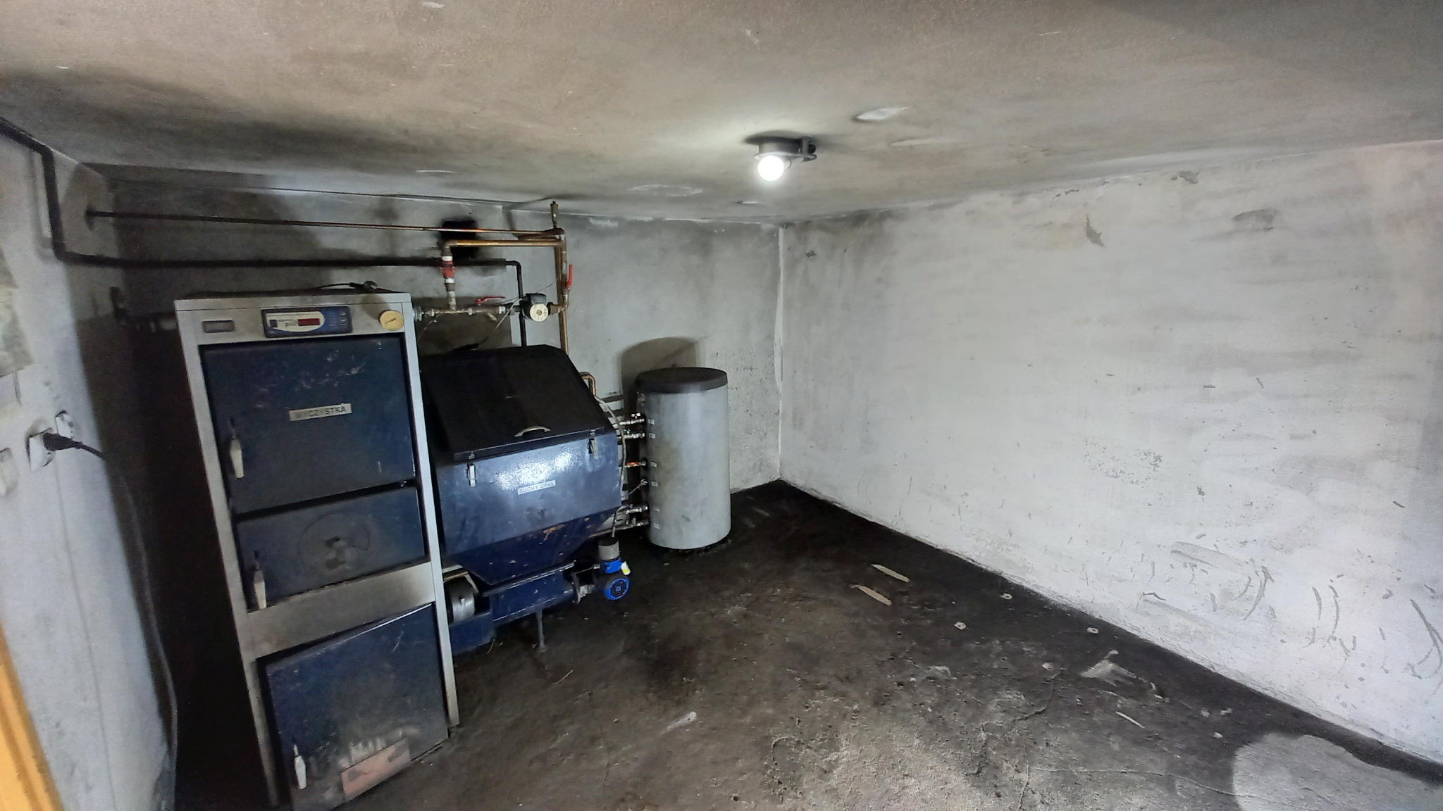 Kotłownia z dwoma piecami na paliwo stałe, jeden z automatycznym podajnikiem, widoczne rury instalacji centralnego ogrzewania, surowe ściany i podłoga.