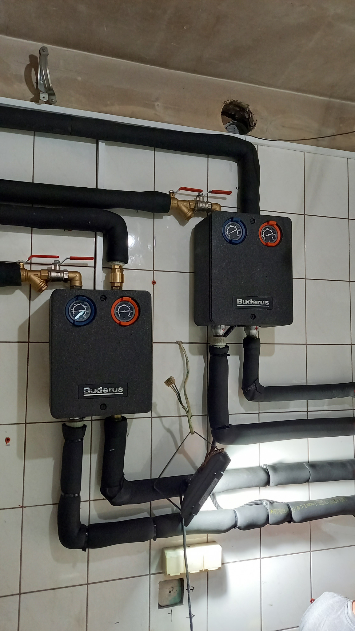 Dwie pompy ciepła Buderus zamontowane na kafelkowanej ścianie, z widocznymi rurami w izolacji i zaworami, instalacja w trakcie podłączenia, widoczny kabel z wtyczką