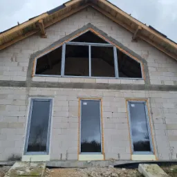 Surowy stan budynku z szarego betonu komórkowego z trzema oknami wstawionymi na parterze i jednym dużym oknem trójkątnym na poddaszu, uszczelnionymi pianką montażową, widok z dołu.