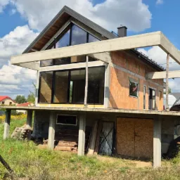 Surowy stan domu z cegły z dużymi oknami i betonowym zadaszeniem, w tle błękitne niebo z chmurami. Widoczne elementy konstrukcyjne i wykończeniowe.