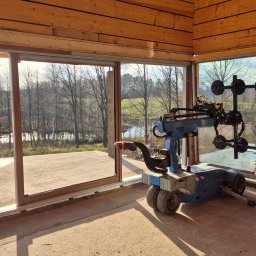 AMS SYSTEMY - Montaż panoramicznego okna w drewnianym domu z widokiem na krajobraz. Użycie specjalistycznego robota do przenoszenia szyb. Praca w toku, widoczny pracownik.