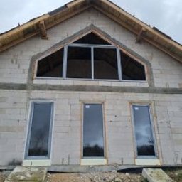 AMS SYSTEMY - Surowy stan budynku z zamontowanymi oknami PCV o grafitowej ramie. Widoczne trzy okna pionowe i jedno trójkątne na poddaszu. Budynek w trakcie budowy.