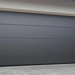 Zamknięta, segmentowa brama garażowa w kolorze grafitowym, z poziomymi przetłoczeniami, oświetlona dwoma zewnętrznymi lampami ściennymi, z elewacją wykończoną kamieniem dekoracyjnym.